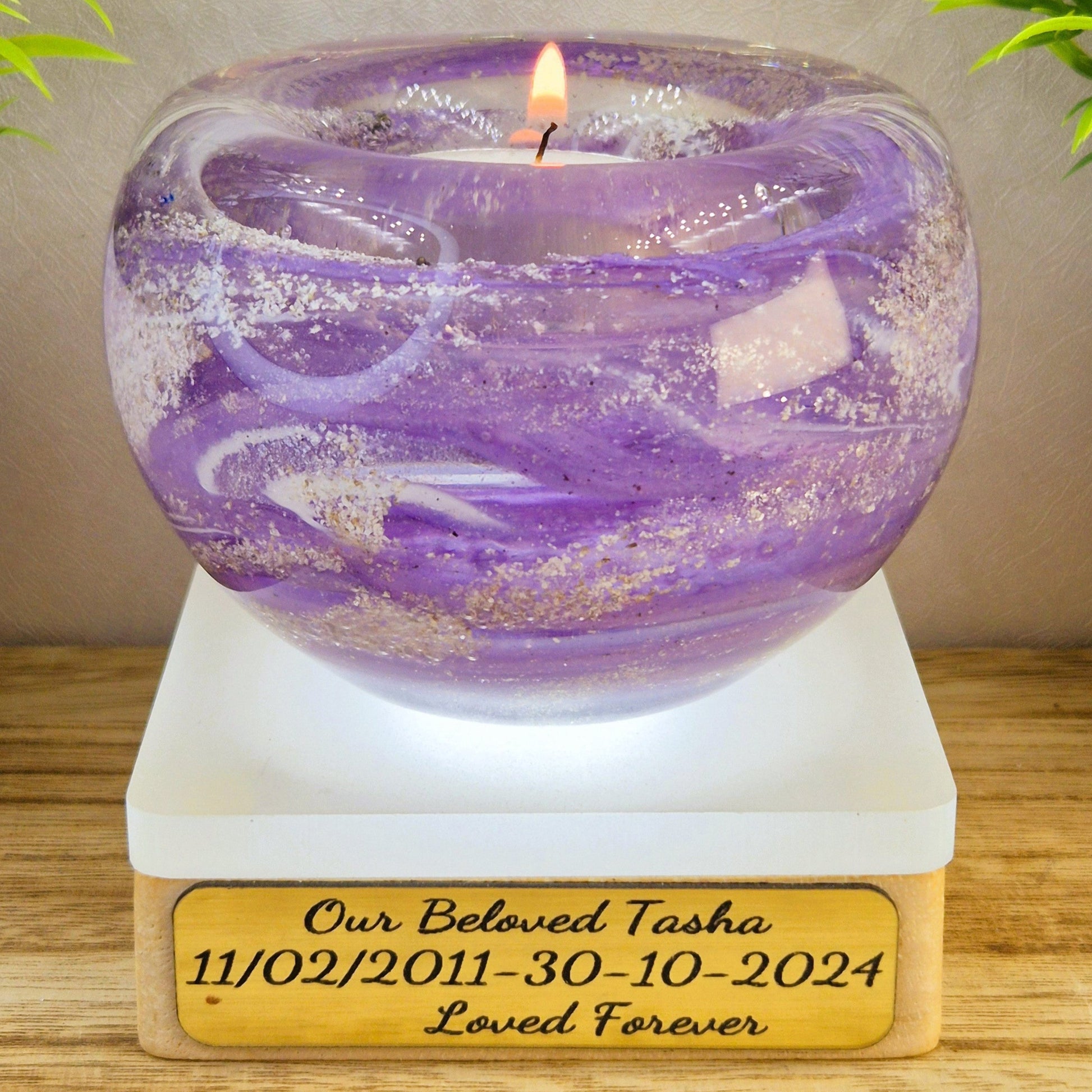Purple candle in a glass holder on a wooden base with engraved text, placed on a wooden surface with plants in the background.